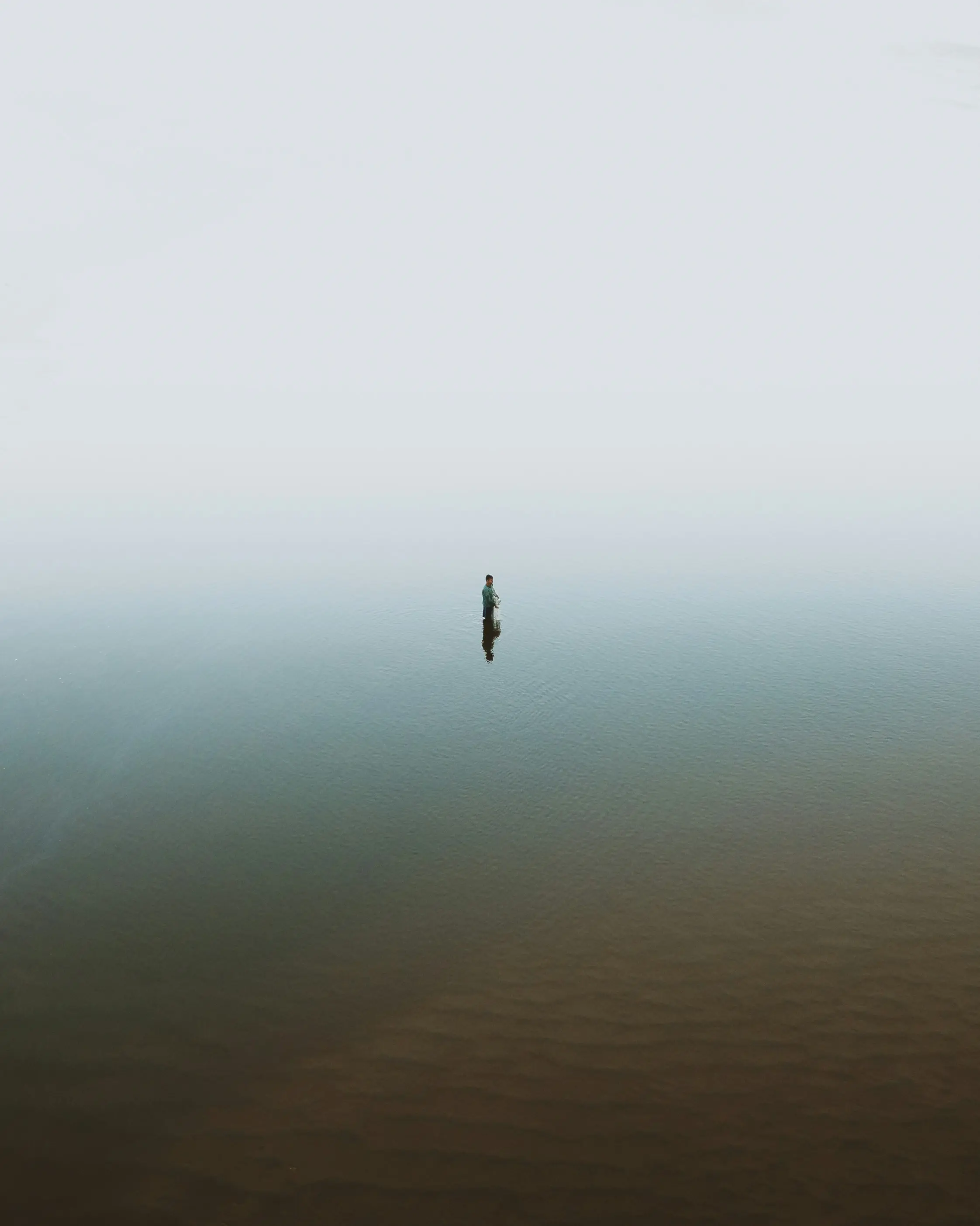 A lone fisherman stands waist-deep in a misty body of water during daytime, surrounded by fog, creating a serene and isolated atmosphere